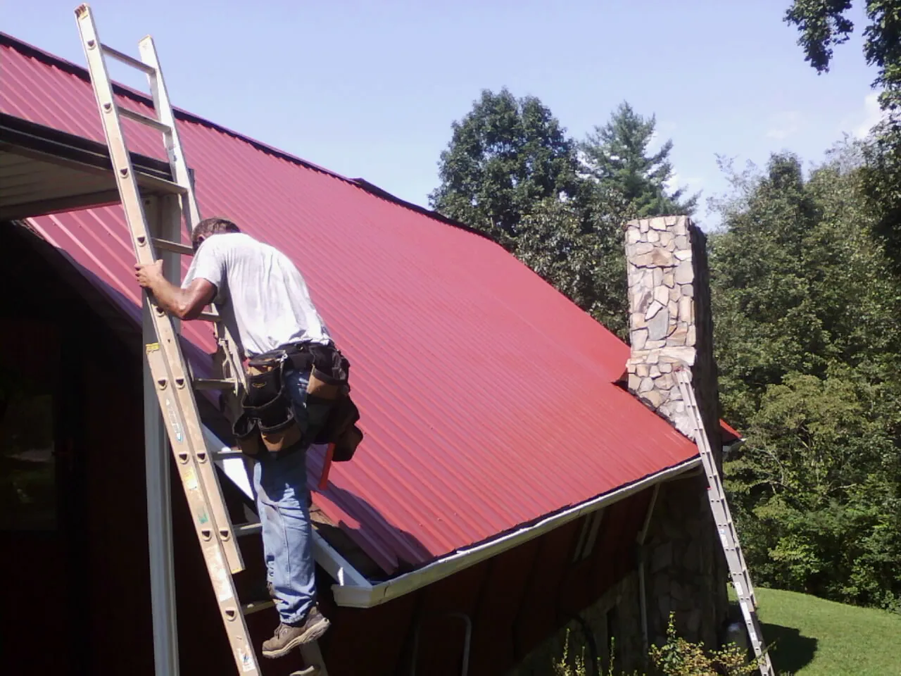 Roofer on a ladder beside a red metal roof and stone chimney — Soffit & Fascia Repair by Roofing Brothers in Fogelsville